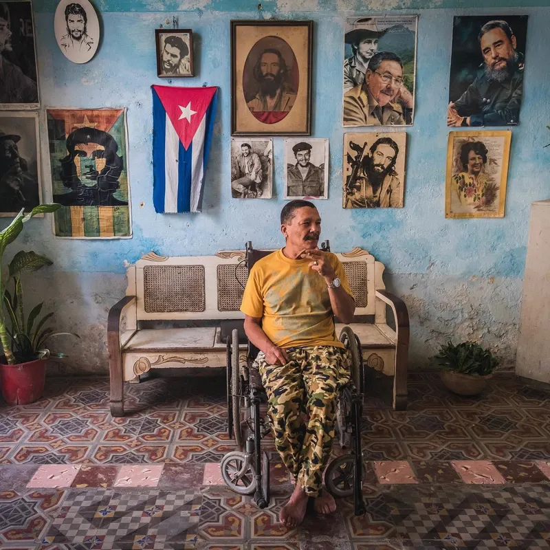 A man in a wheelchair in a tiled living room, the wall behind him covered in framed portraits and a Cuban flag